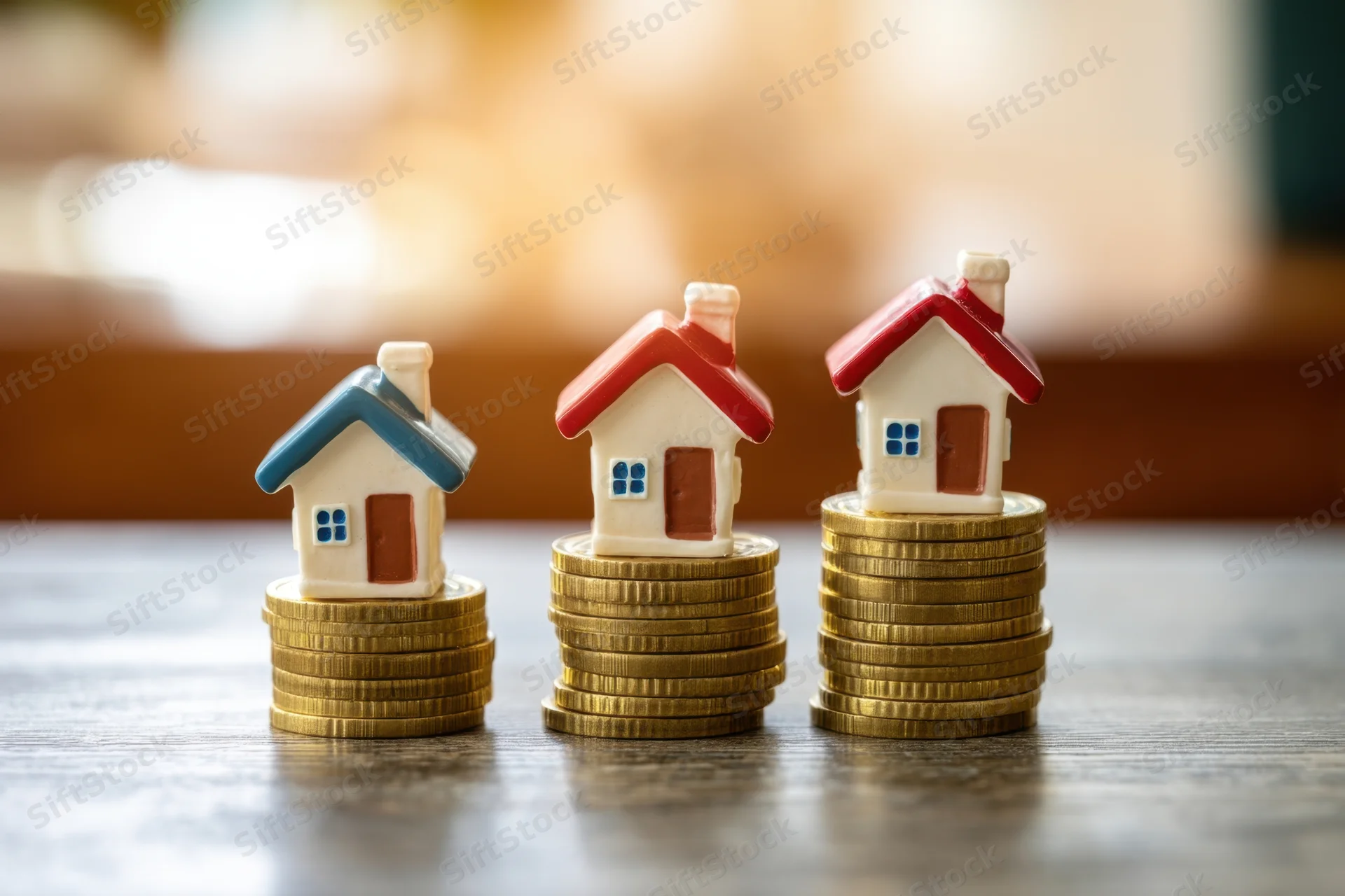 Three miniature houses on stacks of gold coins arranged in a row on a wooden table
