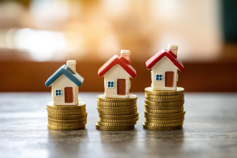 Three miniature houses on stacks of gold coins arranged in a row on a wooden table