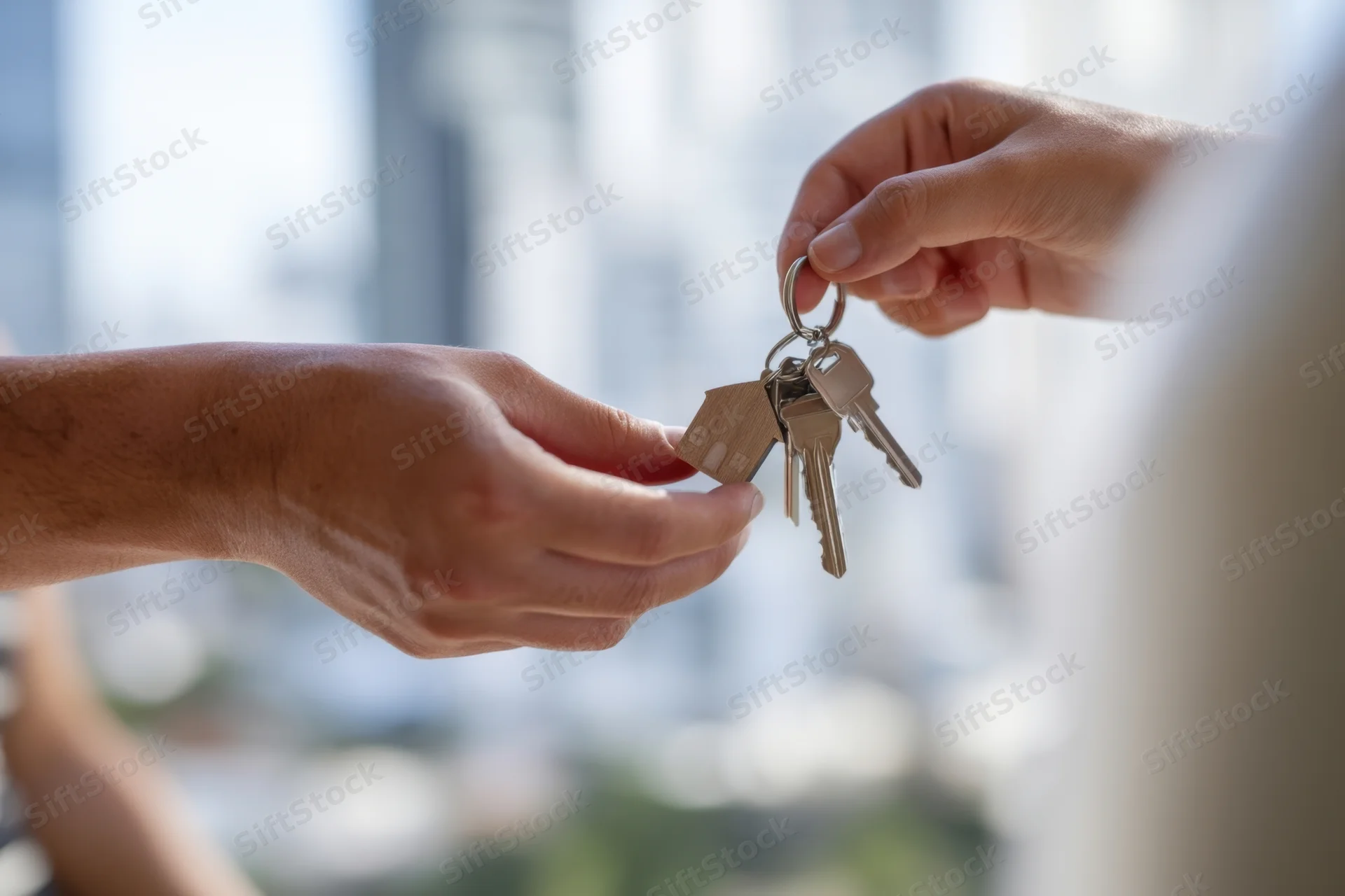 Two hands exchanging keys with a house-shaped keychain against a blurred city view