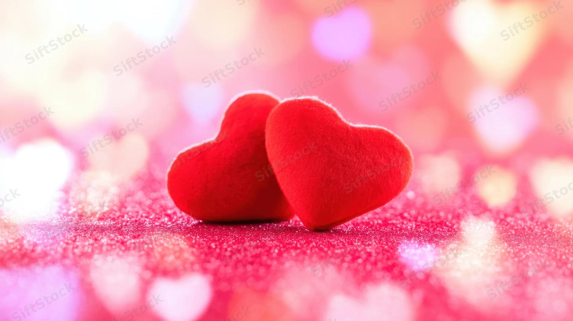 Two red plush hearts resting on glittery pink surface with heart-shaped bokeh lights in background