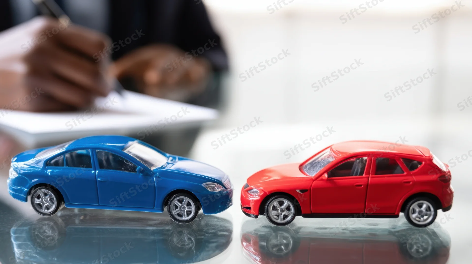 Two toy cars facing each other on a glass desk with a person writing documents in the background