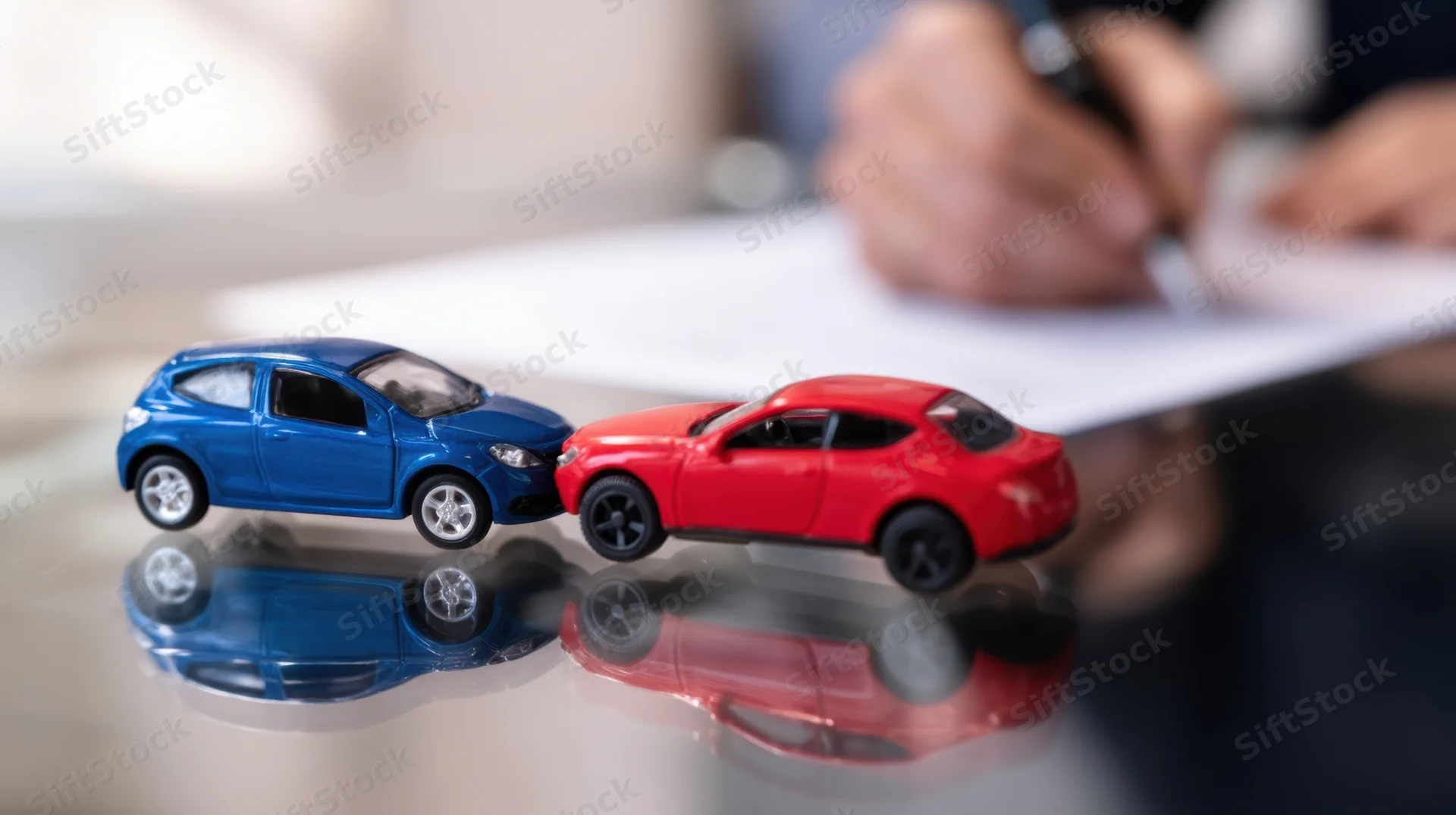Two toy cars colliding on a reflective surface with a hand writing on paper in the background