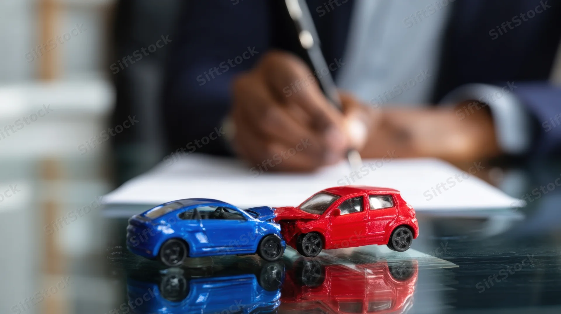 Two toy cars colliding on a desk with a person writing a document in the background