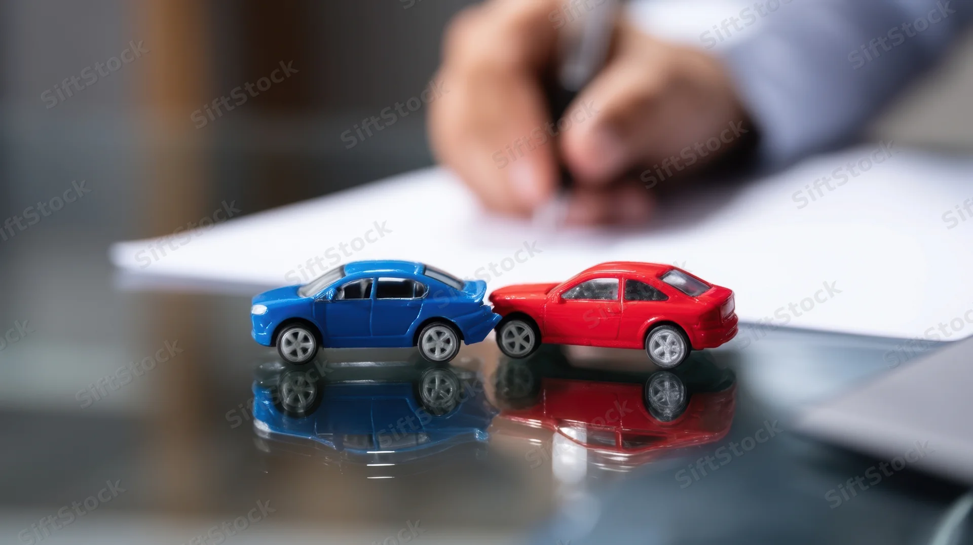 Two toy cars colliding on a reflective desk surface with a hand writing on paper in the background
