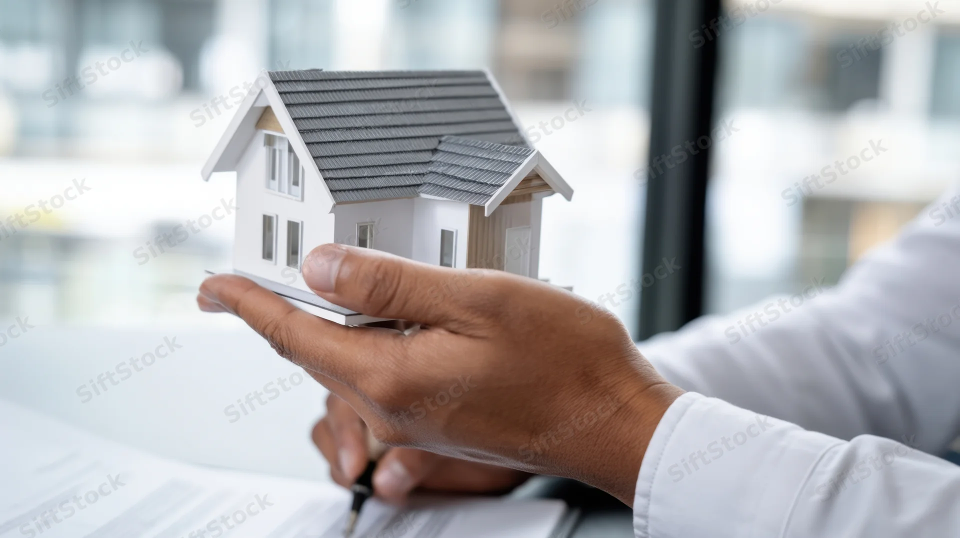 Hand holding a miniature house model while signing a document at a desk in an office