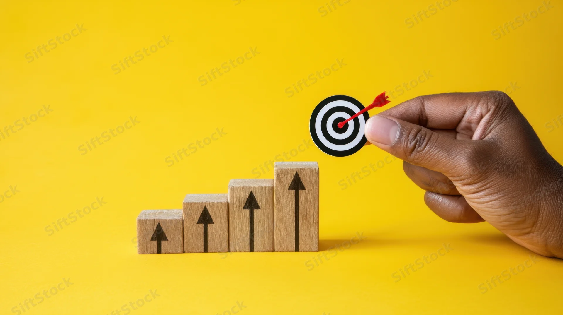 Hand holding target with arrow above wooden blocks arranged in ascending order on yellow background