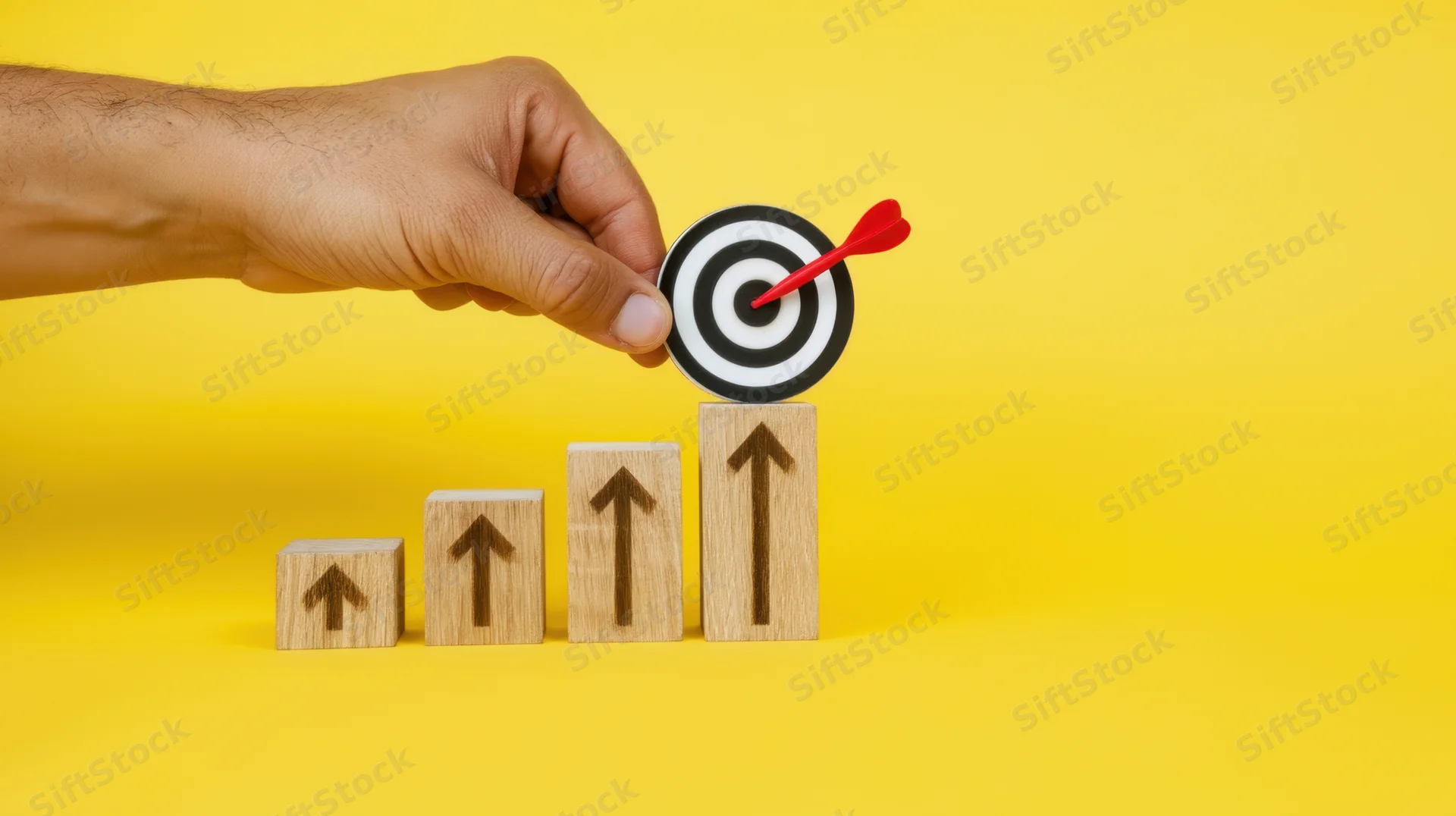 Hand placing a dart in a target atop a row of ascending wooden blocks with arrows, on a yellow background