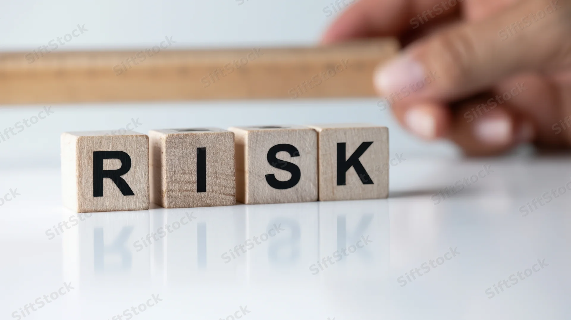 Hand adjusting wooden blocks spelling risk on a reflective white surface with a ruler in the background