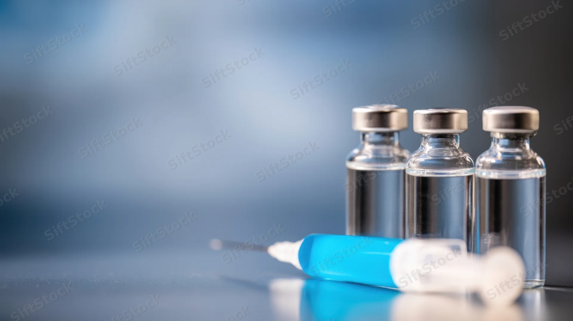 Three glass vaccine vials with a blue syringe on a reflective surface