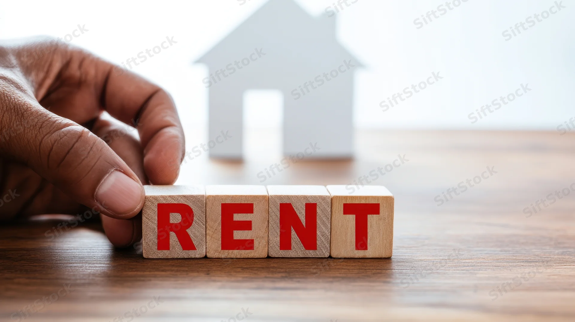 Hand arranging wooden blocks spelling rent on a table with a house silhouette in the background