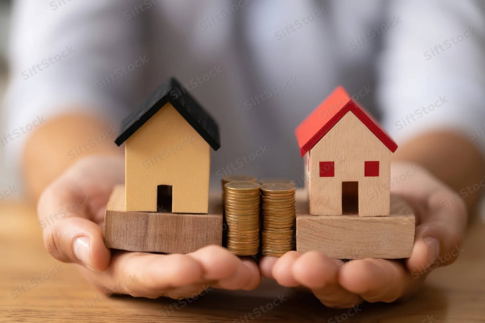 Hands holding wooden house models and stacks of coins on a table