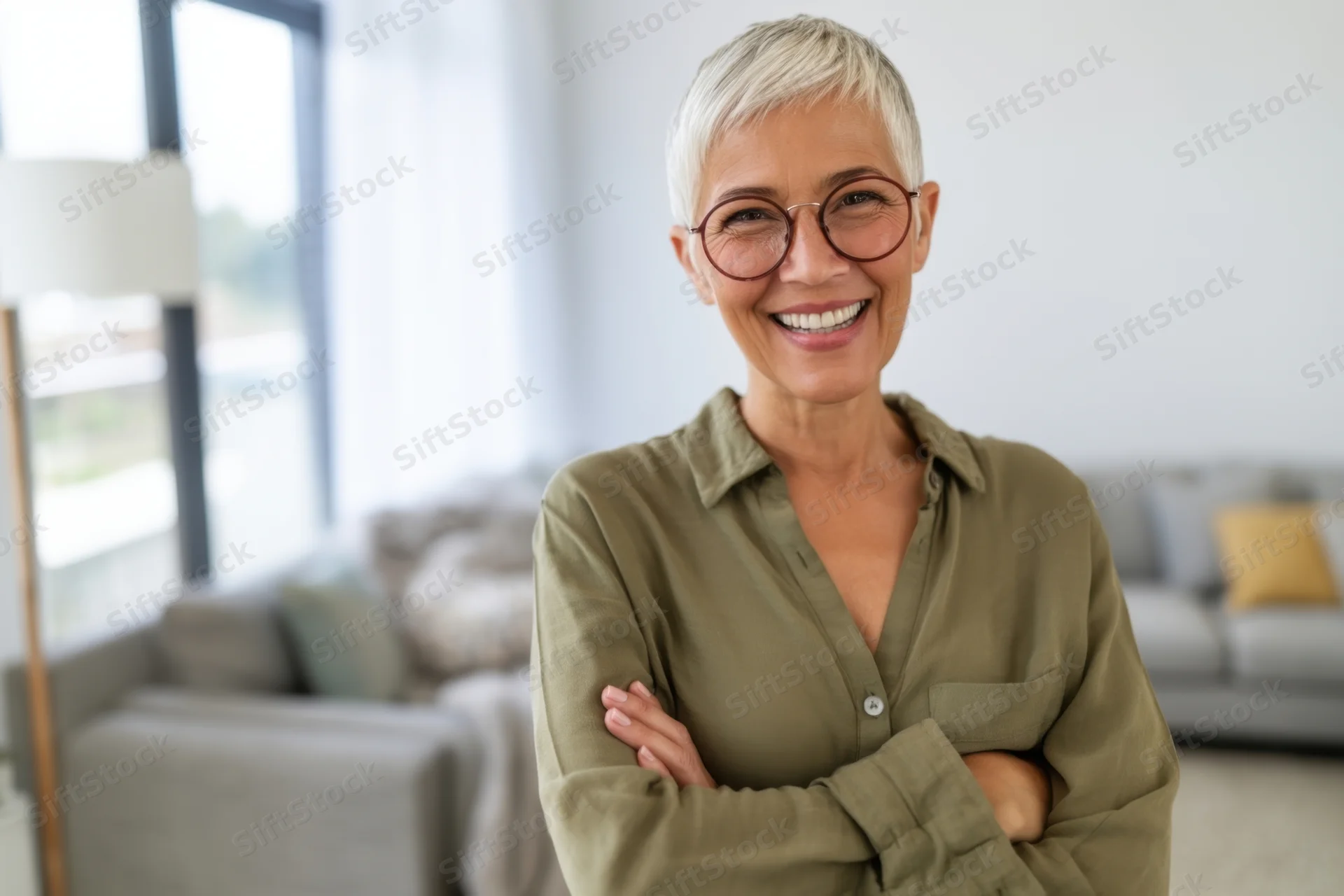 Smiling senior woman with short silver hair and round glasses standing with arms crossed in a modern living room