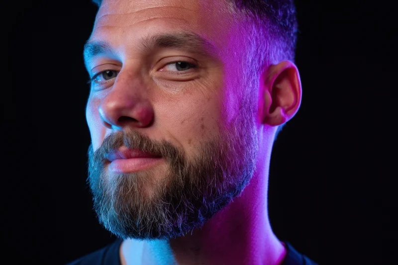 Close-up portrait of a man with a neatly trimmed beard under blue and purple neon lighting against a black background