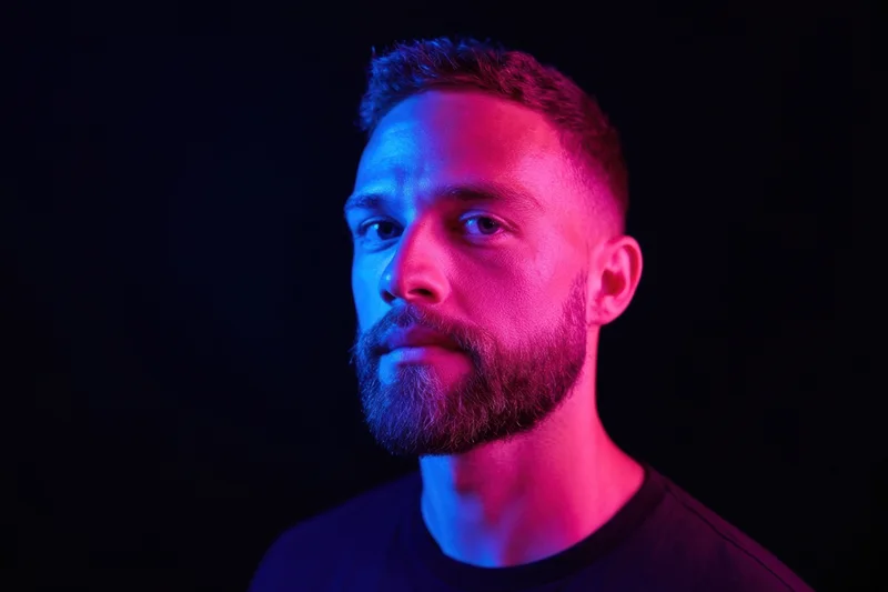 Close-up portrait of a man with a neatly trimmed beard under blue and pink neon lighting in a dark studio