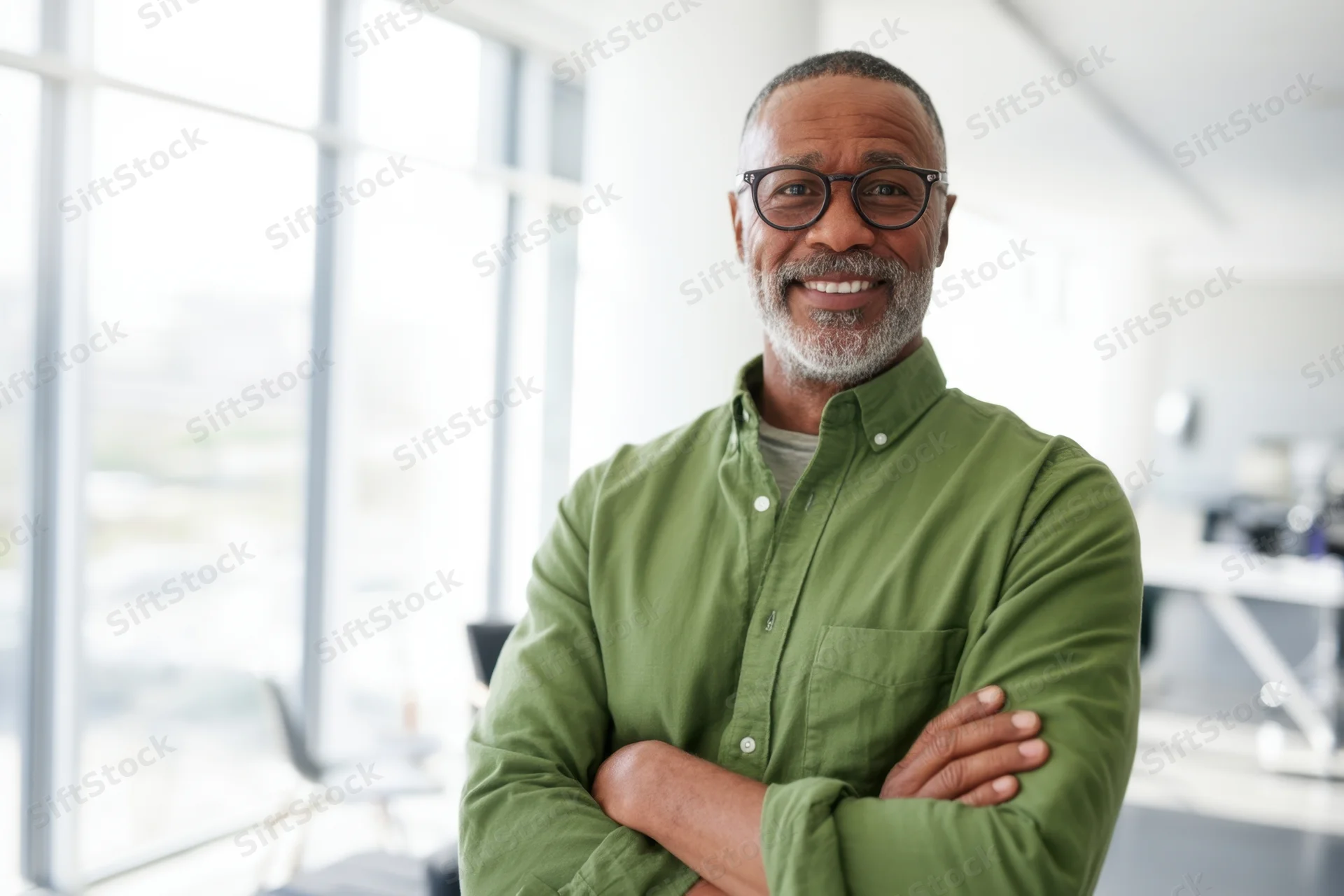 Confident middle-aged man with glasses smiling with arms crossed in bright modern office interior