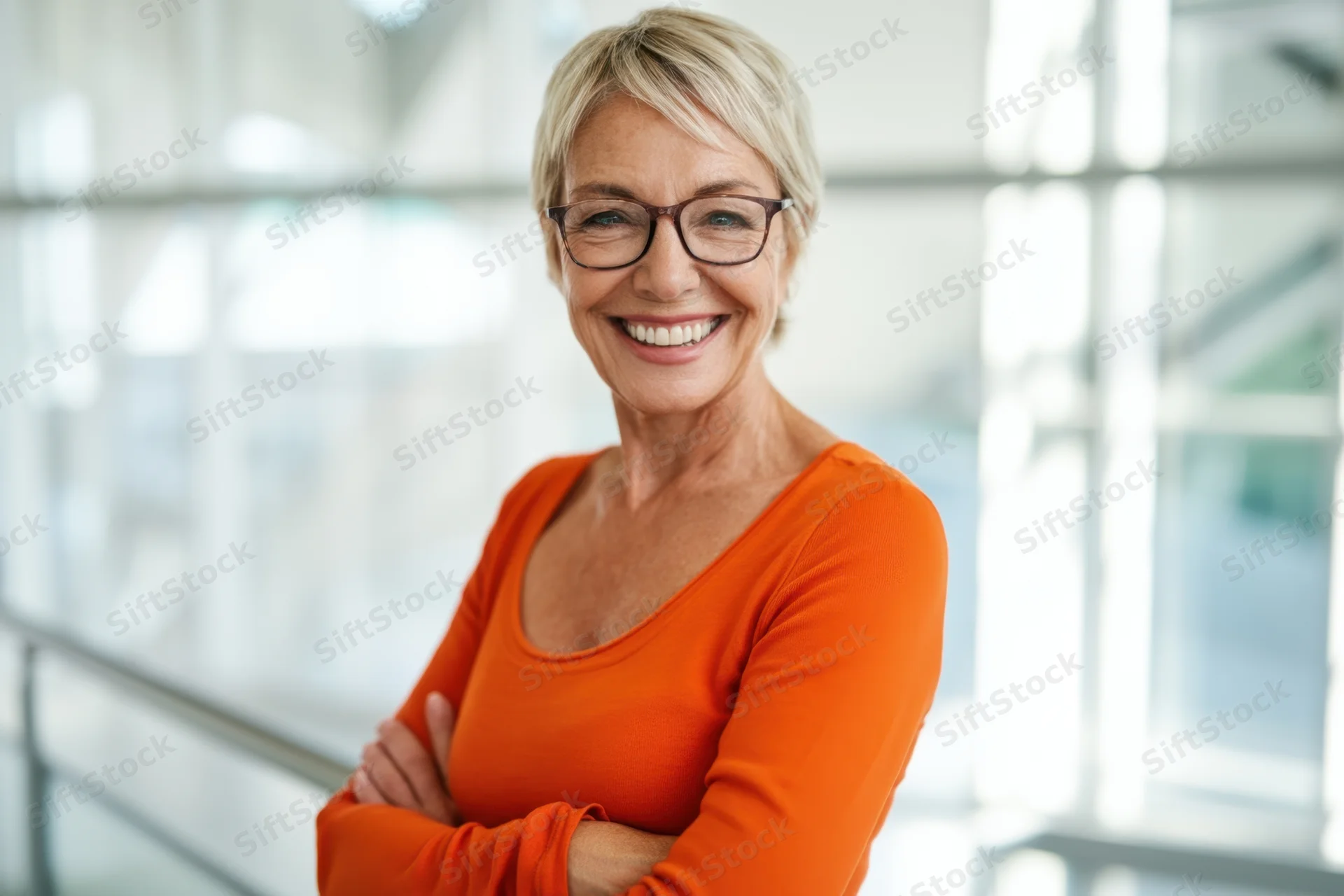 Confident senior woman with short blonde hair and glasses smiling with arms crossed in a modern office interior