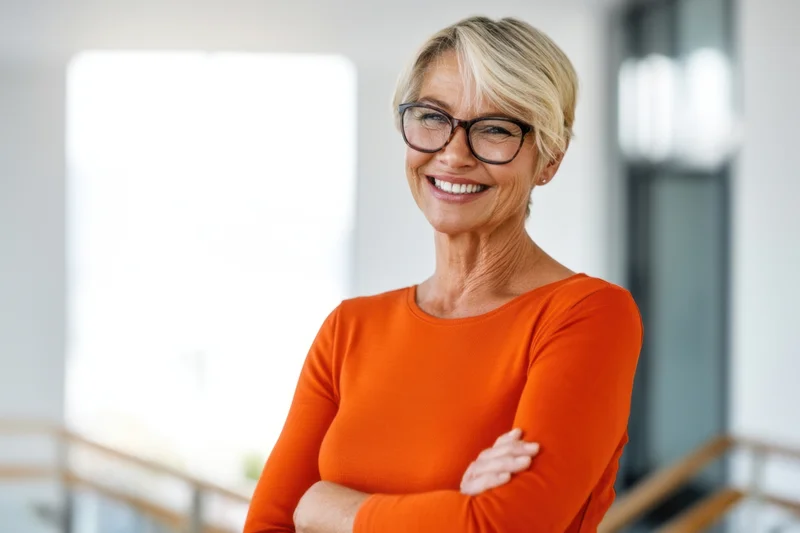 Confident woman with short blonde hair and glasses smiling with arms crossed in a modern office