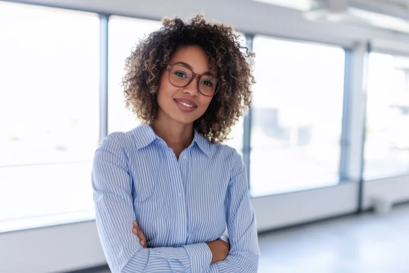 Confident woman with curly hair and glasses smiling with arms crossed in a modern office space