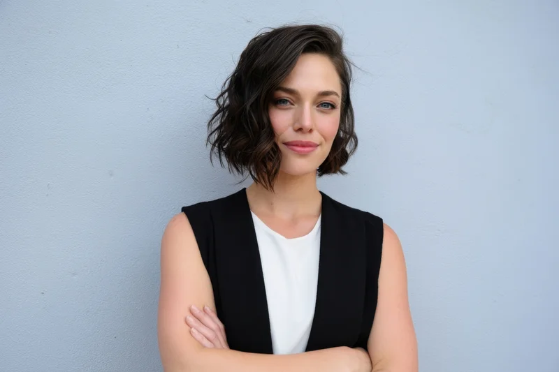 Confident woman with short wavy dark hair standing against a light blue wall with arms crossed