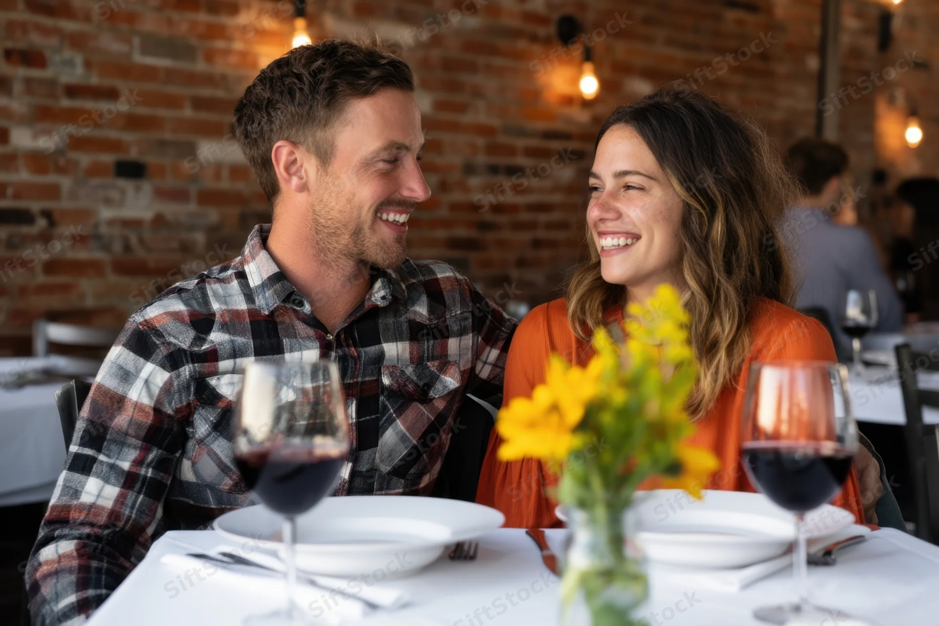 Couple smiling at each other during dinner at a brick-walled restaurant table with wine and flowers