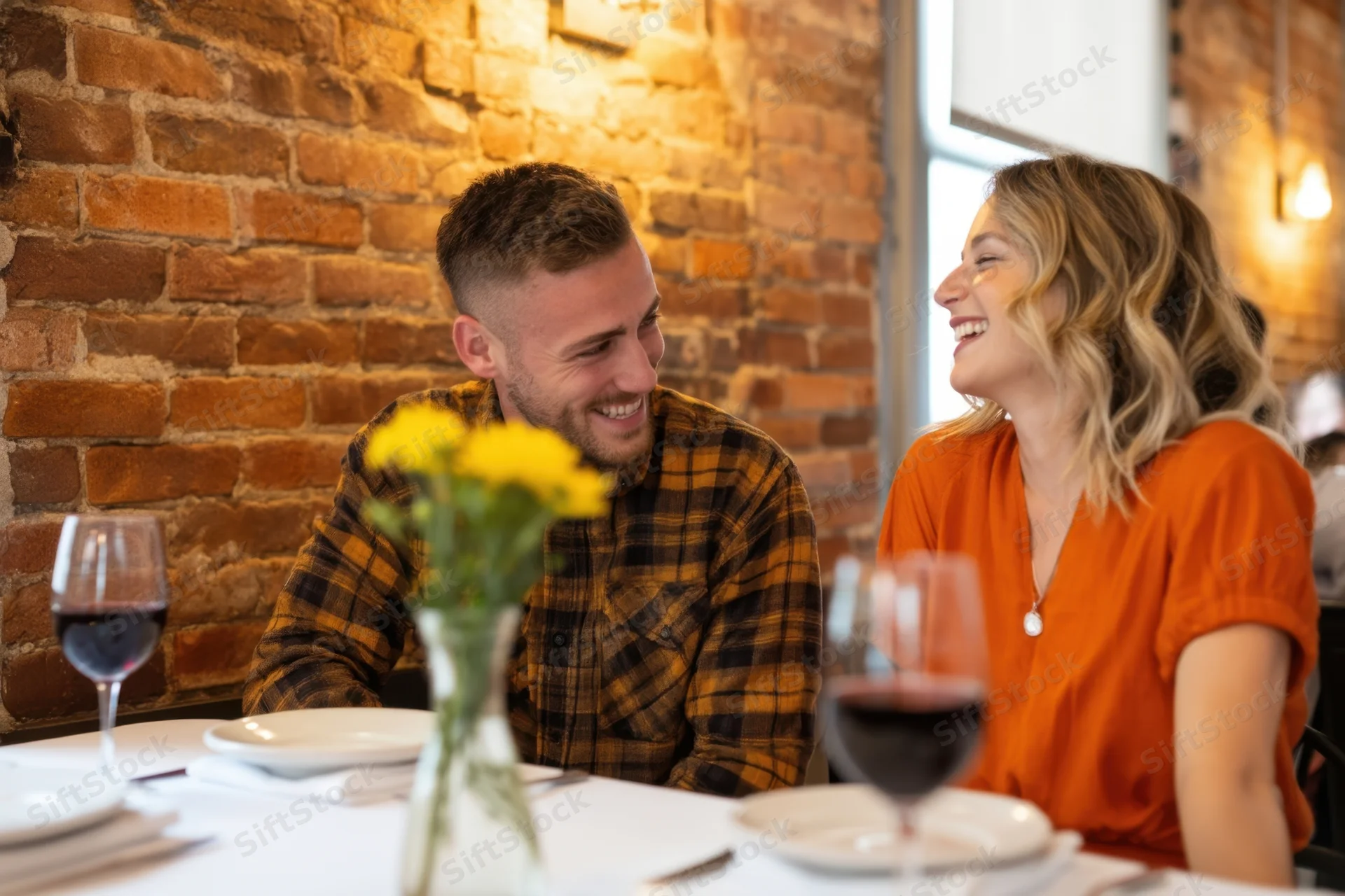 Couple smiling and laughing at dinner table in brick-walled restaurant with wine and flowers