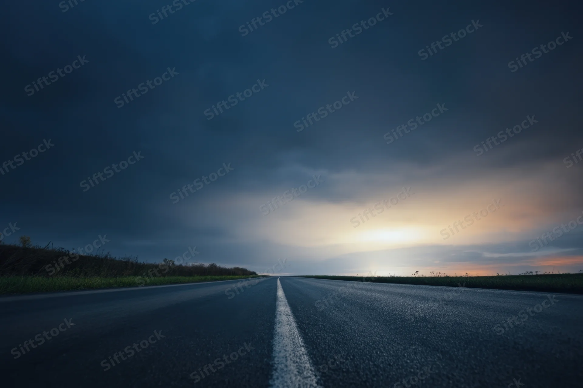 Straight asphalt road under dark stormy sky with distant sunset glow at twilight