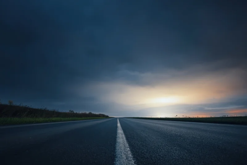 Straight asphalt road under dark stormy sky with distant sunset glow at twilight