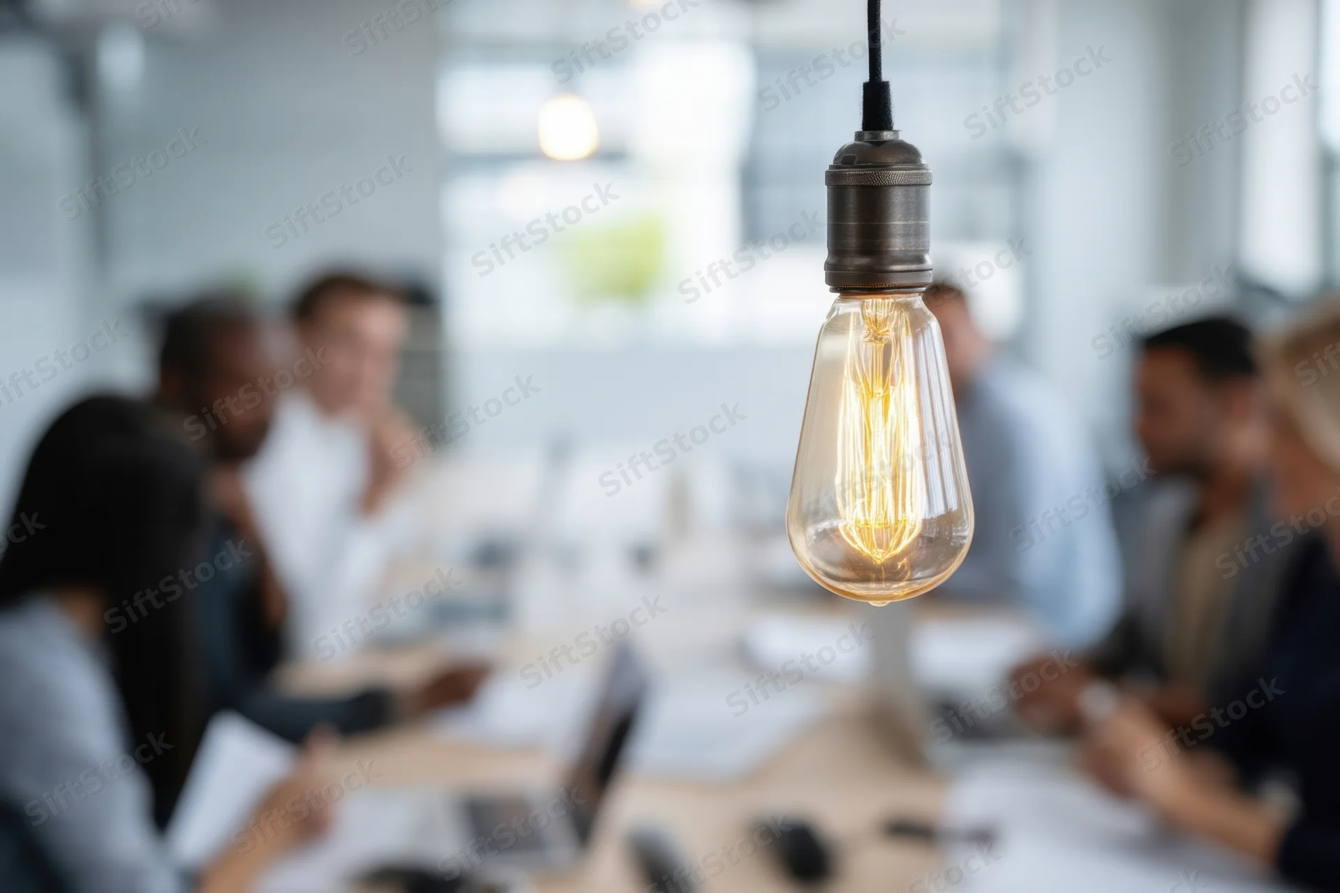 Glowing lightbulb hanging above a diverse group of professionals in a conference meeting at a long table in a modern office