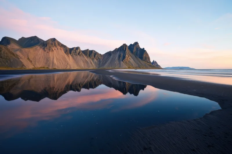 Jagged mountain peaks reflected in still water along a black sand beach at sunset