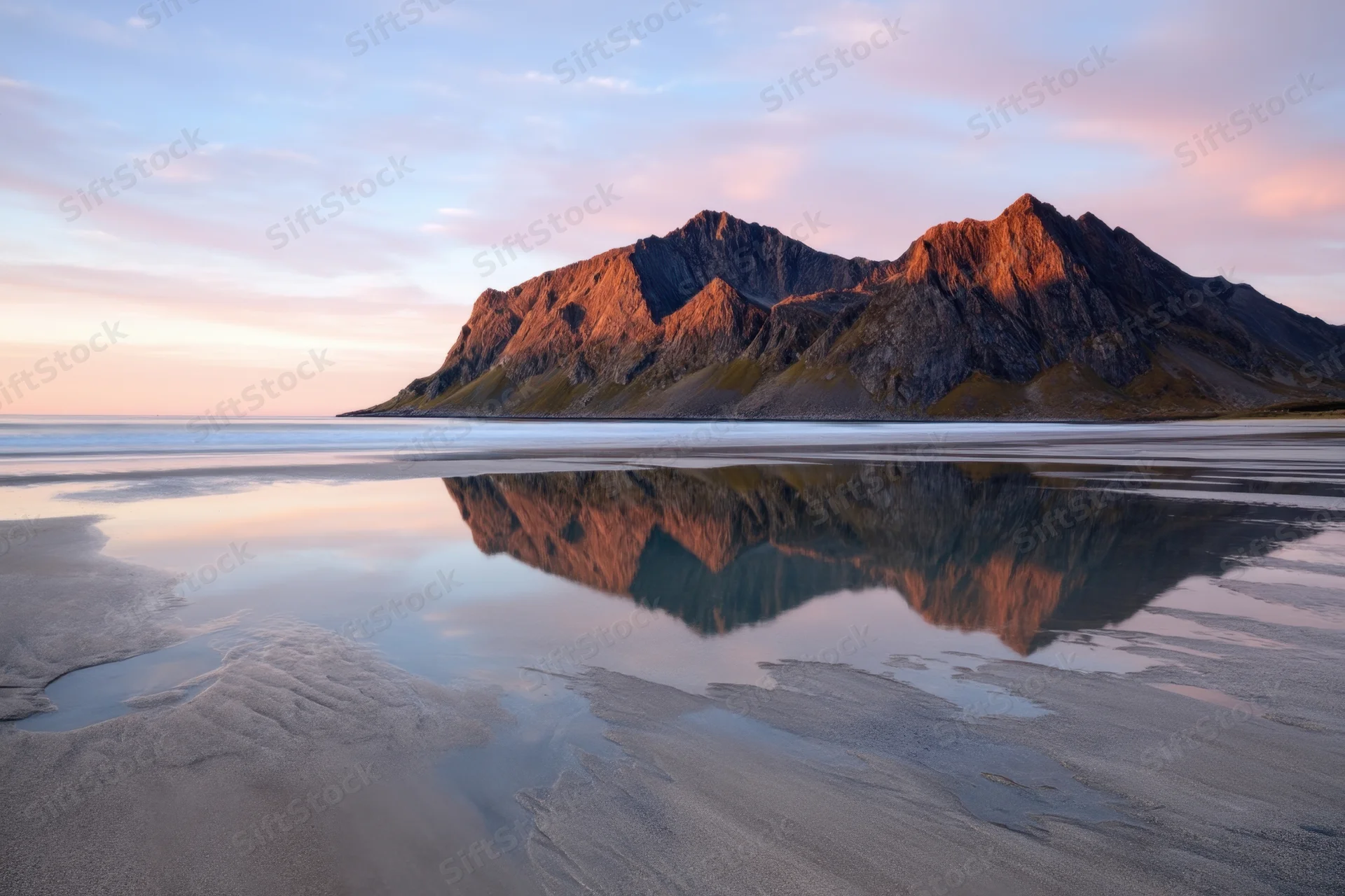 Jagged mountain peaks reflected in calm water along a sandy beach at sunset