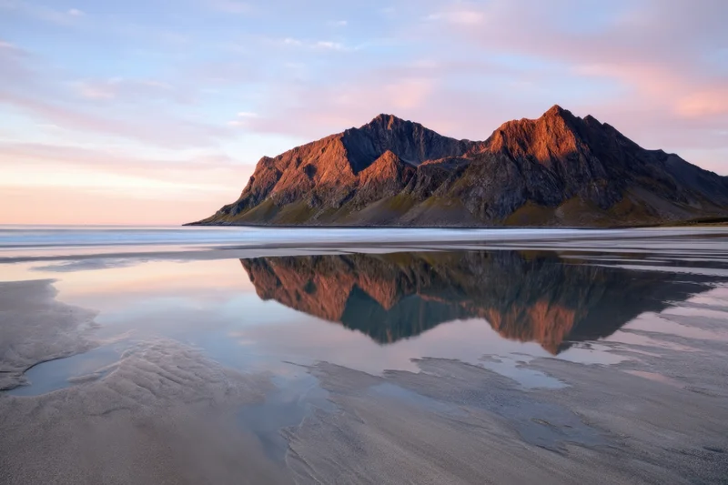 Jagged mountain peaks reflected in calm water along a sandy beach at sunset