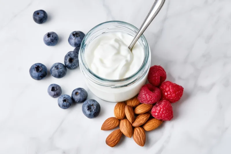 Glass jar of creamy yogurt with spoon surrounded by blueberries, raspberries and almonds on marble surface