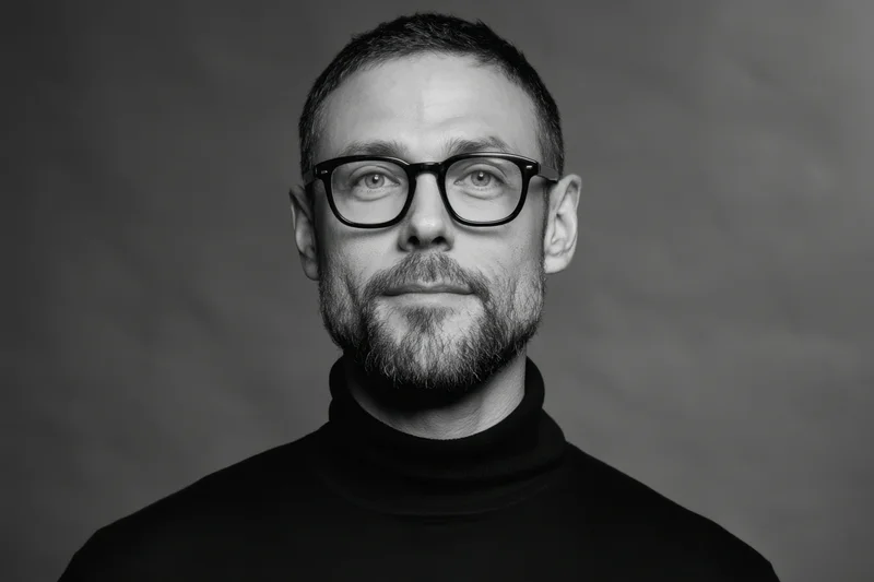 Man with short dark hair and beard wearing black turtleneck and glasses in studio portrait