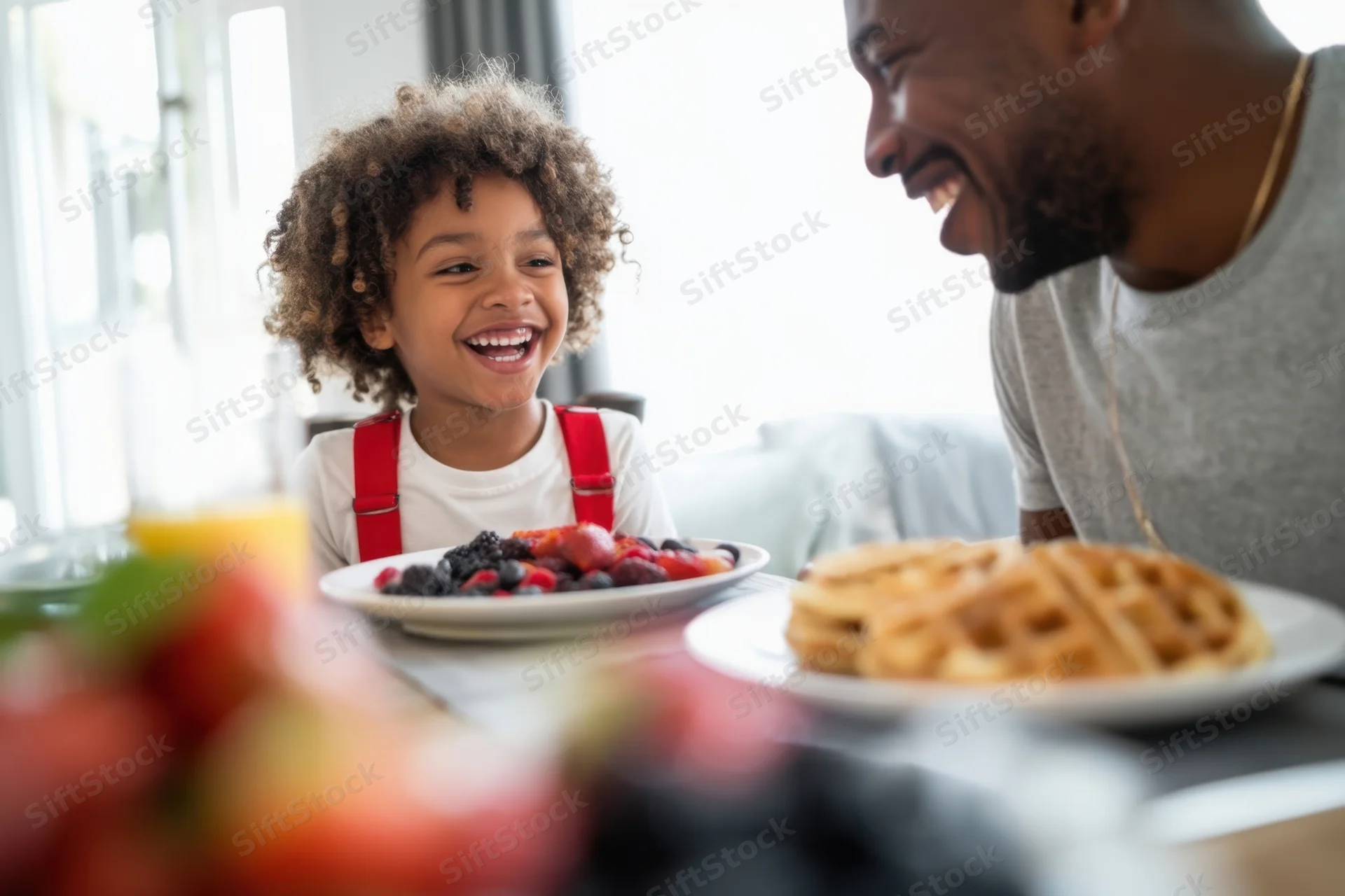 Child laughing at breakfast table with father, plates of waffles and fruit in front of them