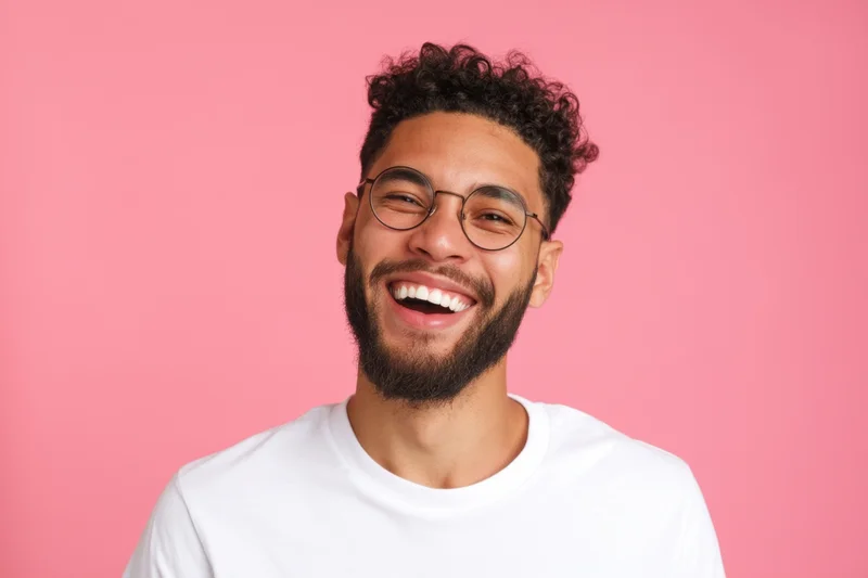 Young man laughing with curly hair and round glasses against pink background wearing white t-shirt