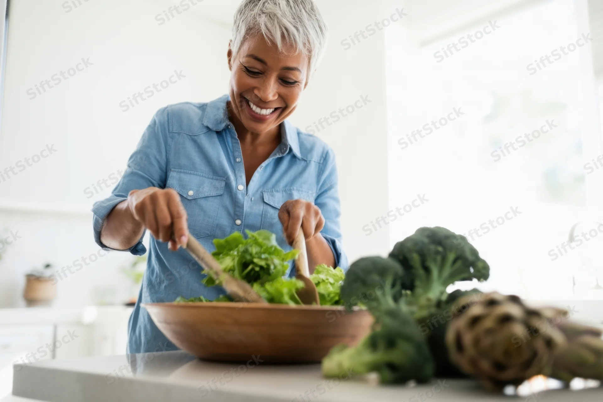 Mature woman smiling while tossing salad in wooden bowl in kitchen