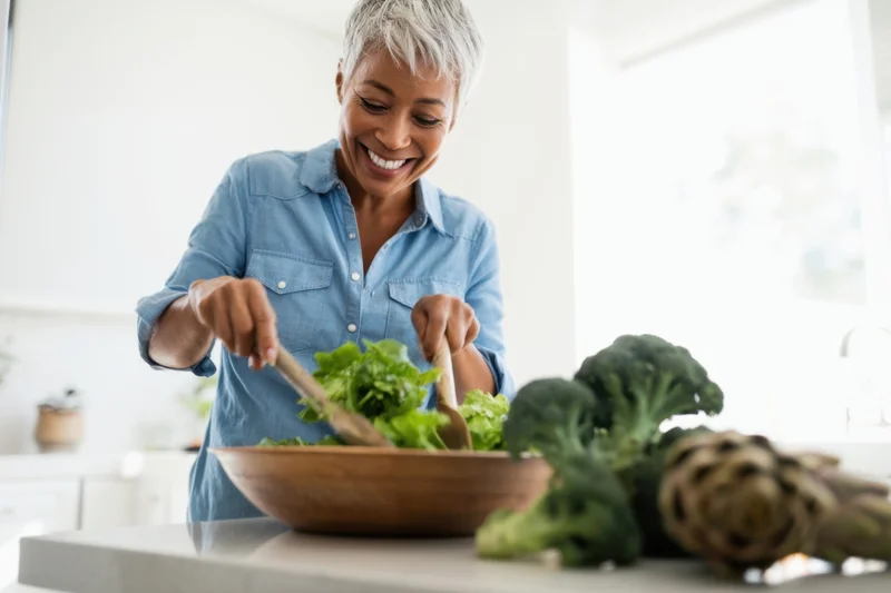Mature woman smiling while tossing salad in wooden bowl in kitchen