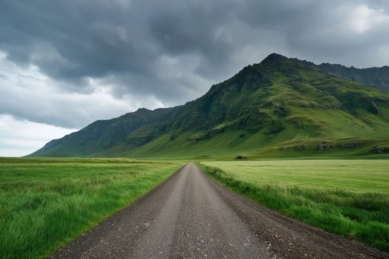 Gravel road leading through green fields toward a grassy mountain under cloudy sky