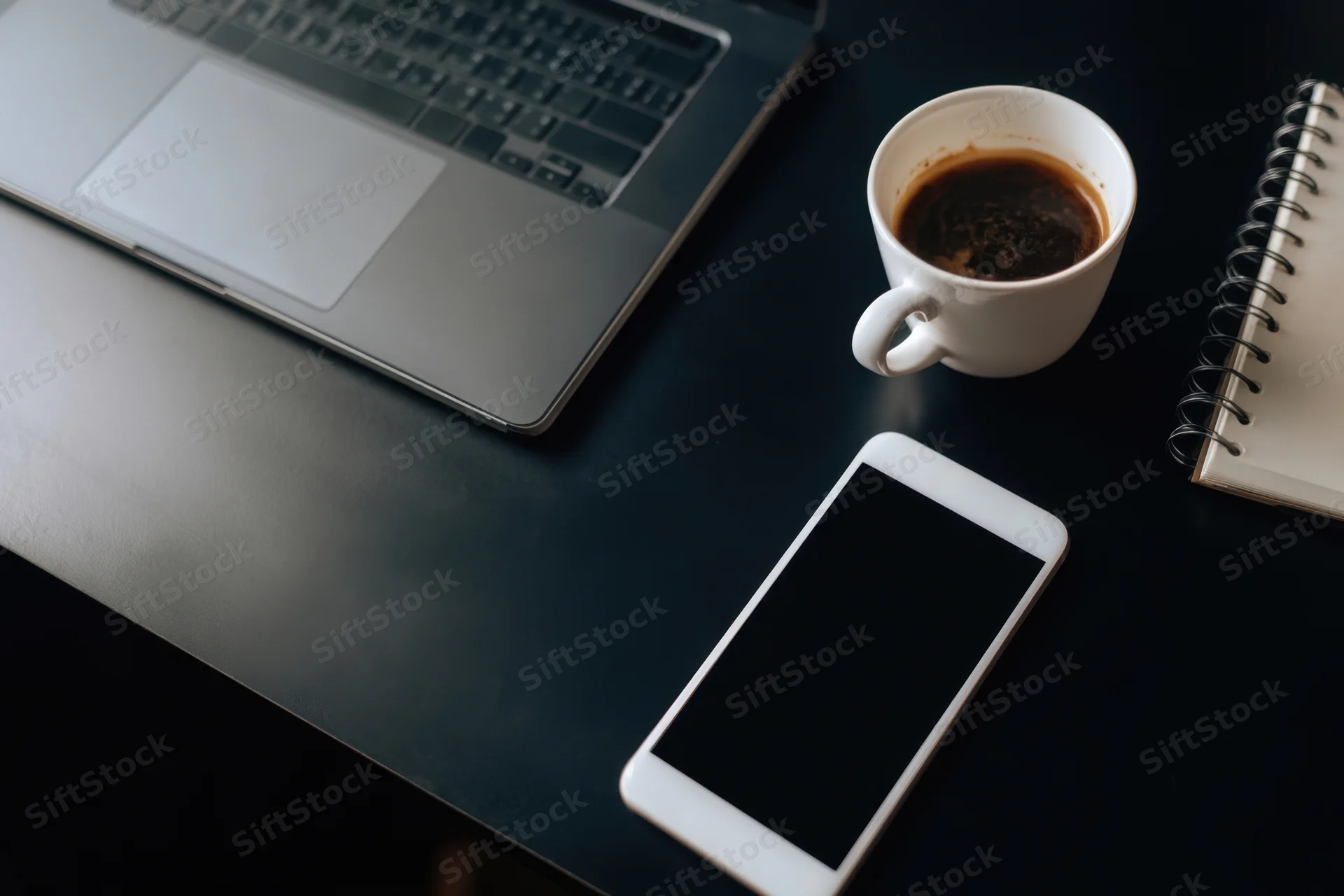 Dark desk with laptop, coffee mug, smartphone and notebook arranged for work