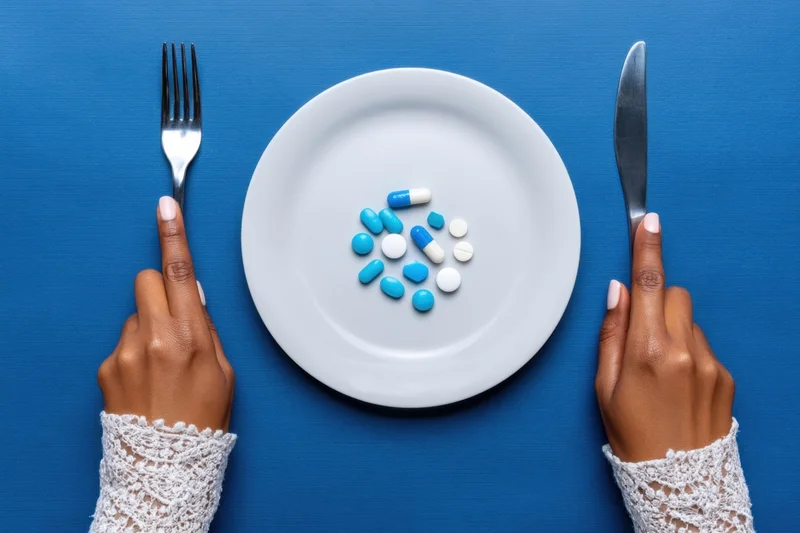 Woman's hands holding fork and knife framing a plate with pills on a blue surface
