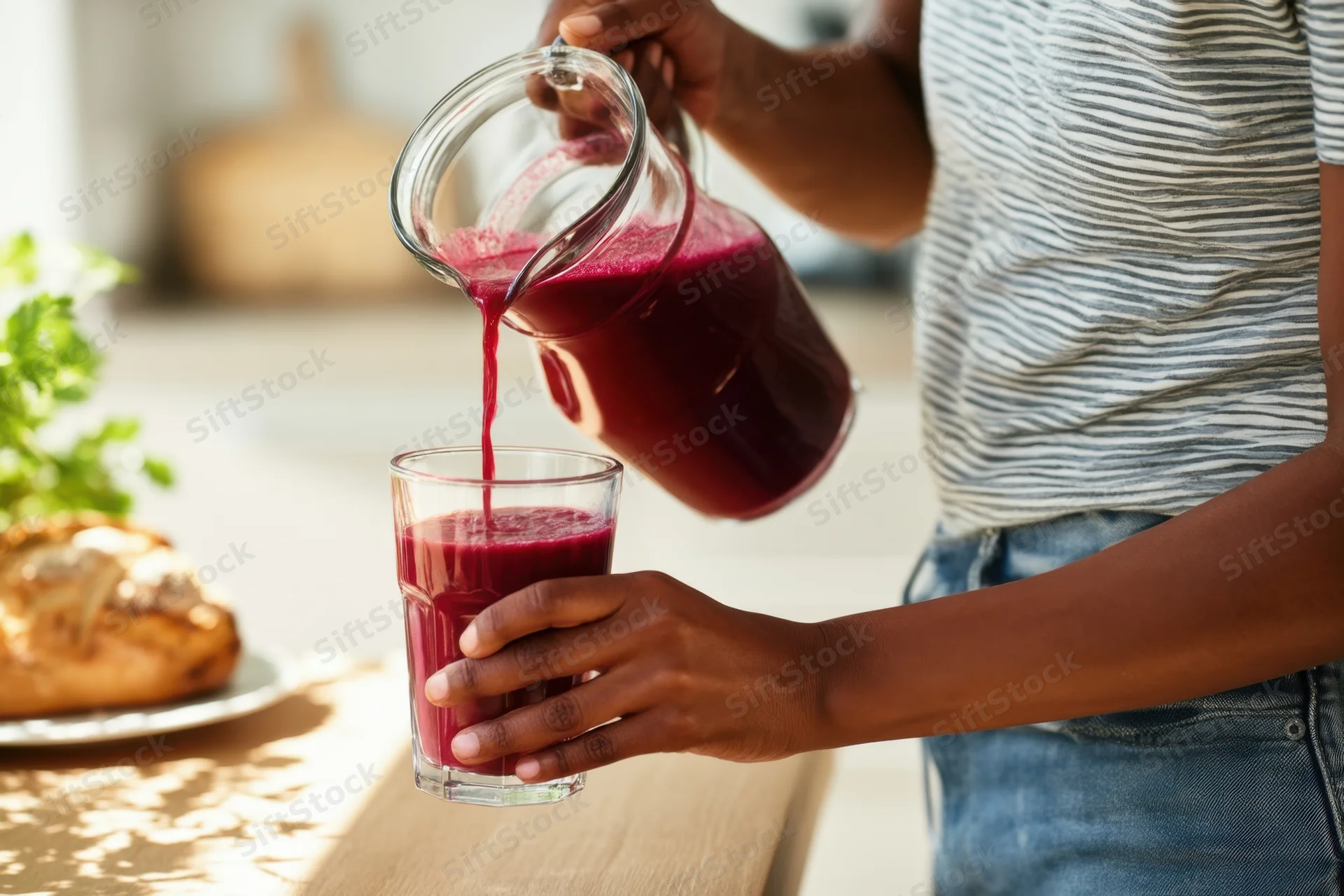 Woman pouring red juice from a glass pitcher into a glass at a kitchen table during breakfast