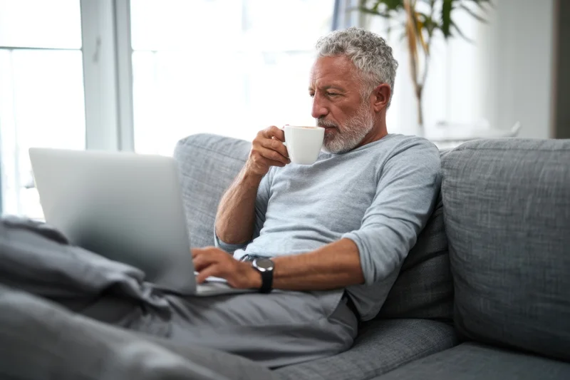 Senior man with gray beard sipping coffee while using laptop on gray sofa in living room