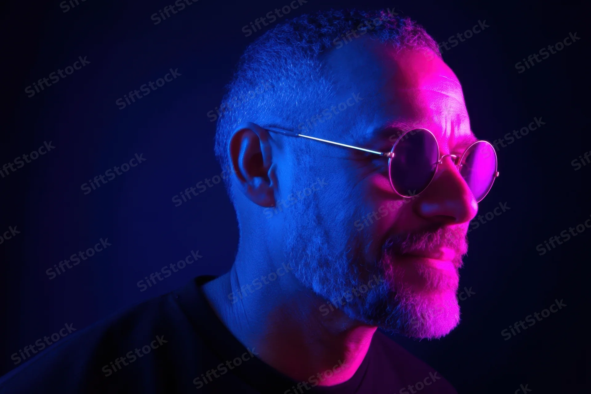 Side-profile portrait of a man with round sunglasses and a beard under blue and pink neon lighting in a dark studio