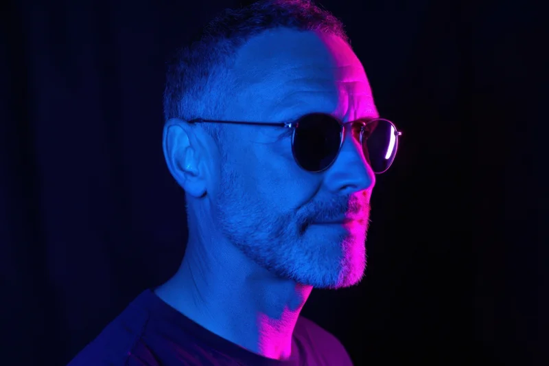 Man in round sunglasses in profile under blue and purple neon lighting against dark background