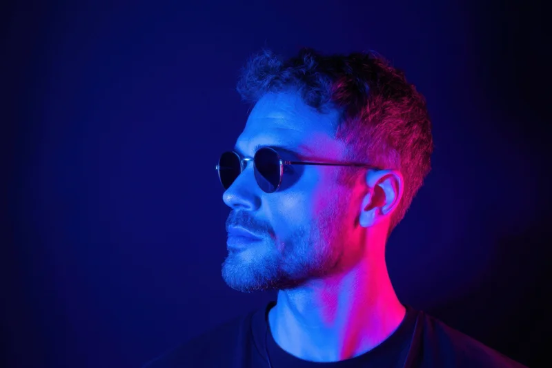 Man in round sunglasses in profile under blue and pink neon lighting in studio