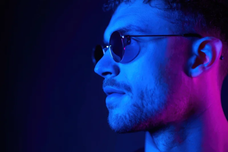 Side profile of a man with round sunglasses under blue and pink neon lighting