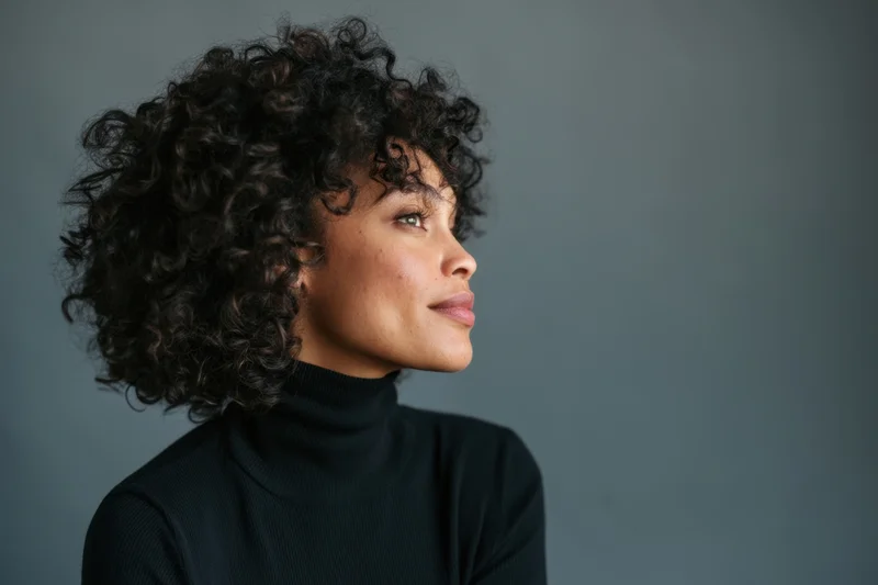 Woman with natural curly hair in profile wearing black turtleneck against gray background