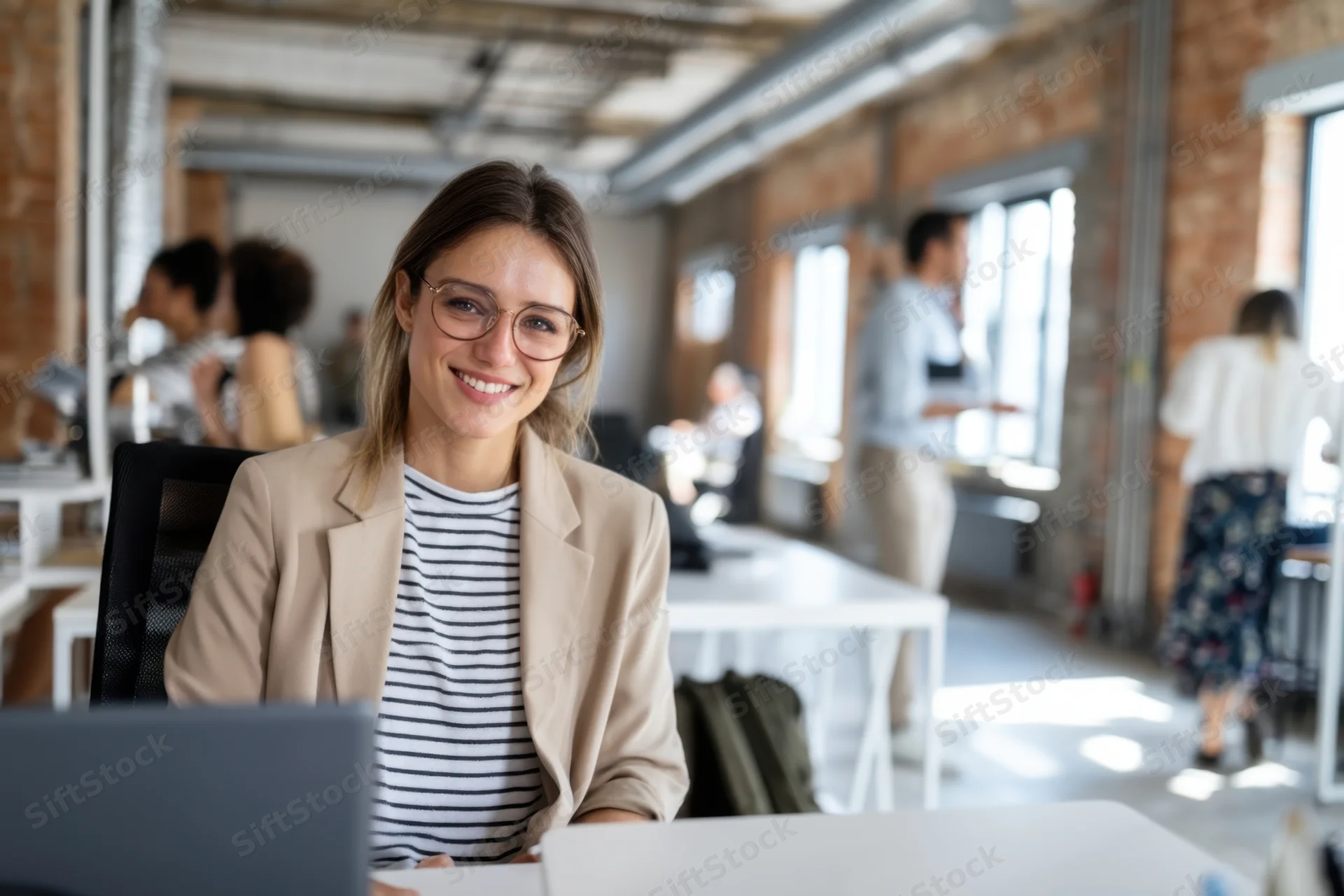 Smiling woman in beige blazer and striped shirt sitting at desk in modern open-plan office with colleagues working in background