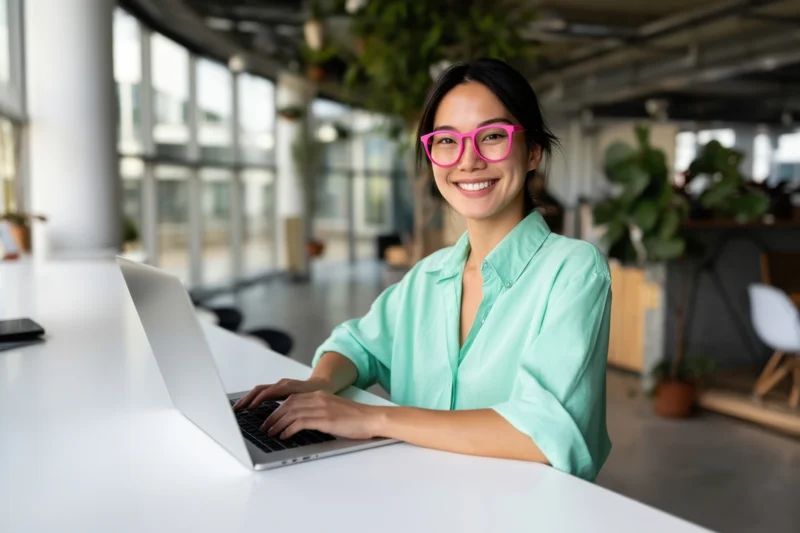 Smiling asian woman typing on laptop at modern office desk