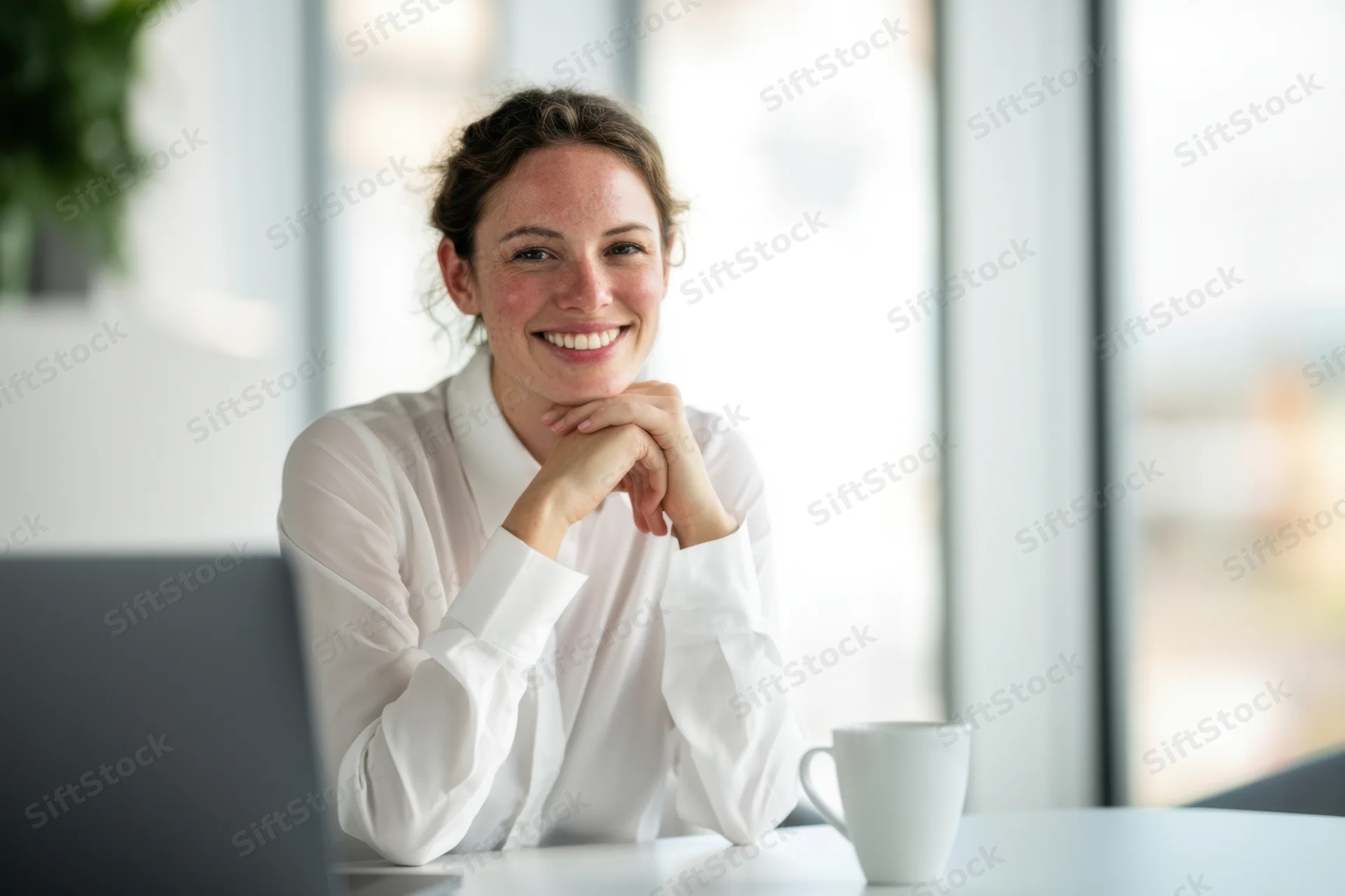 Smiling woman sitting at office desk with laptop and mug by large windows